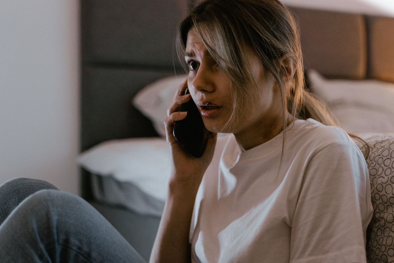 A worried woman sitting on the edge of a bed with a phone pressed to her ear.